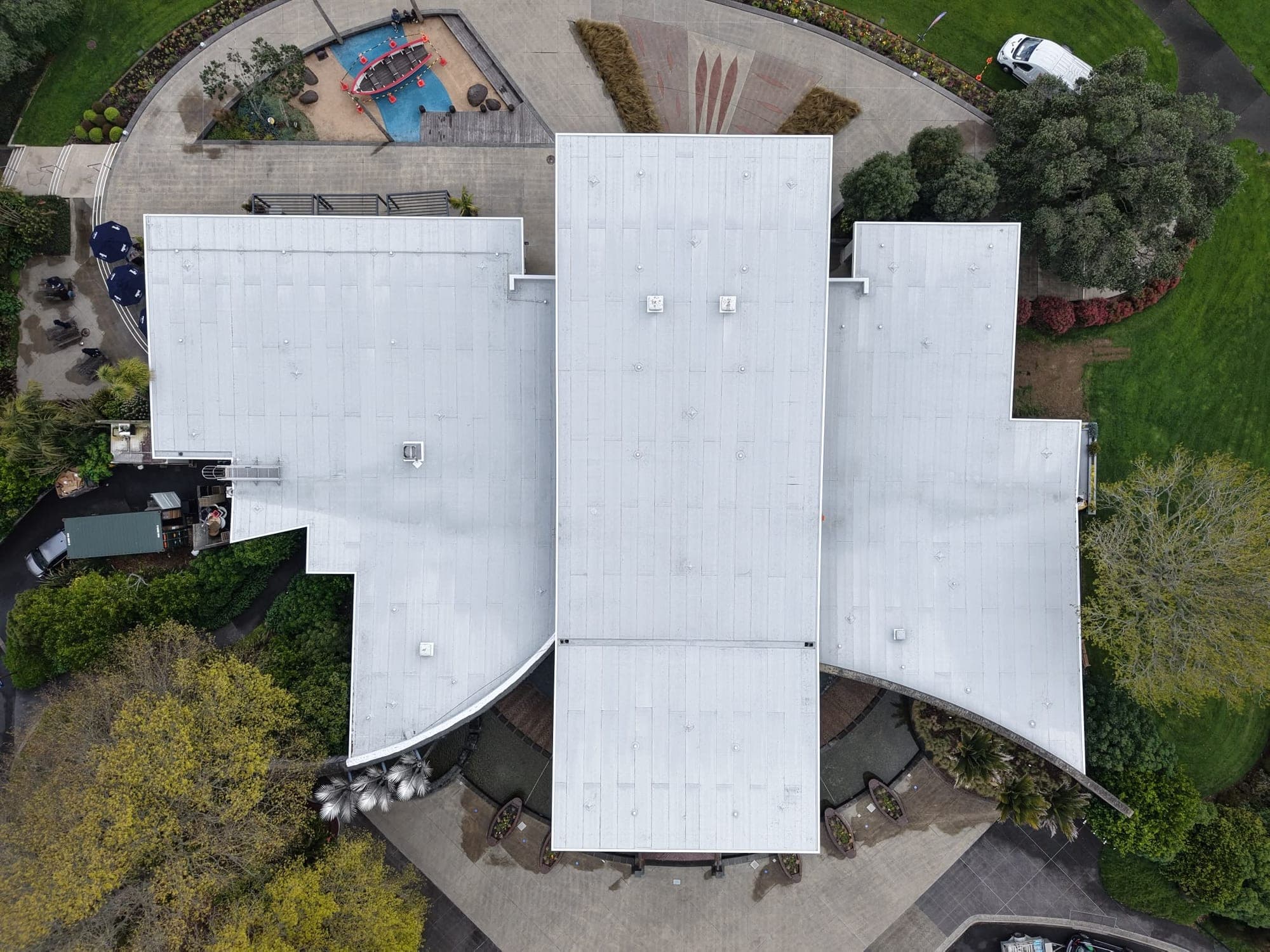 Drone view looking straight down on the finished white membrane roof of the Auckland Botanic Gardens Visitors Centre, with gardens, trees and the forecourt around it