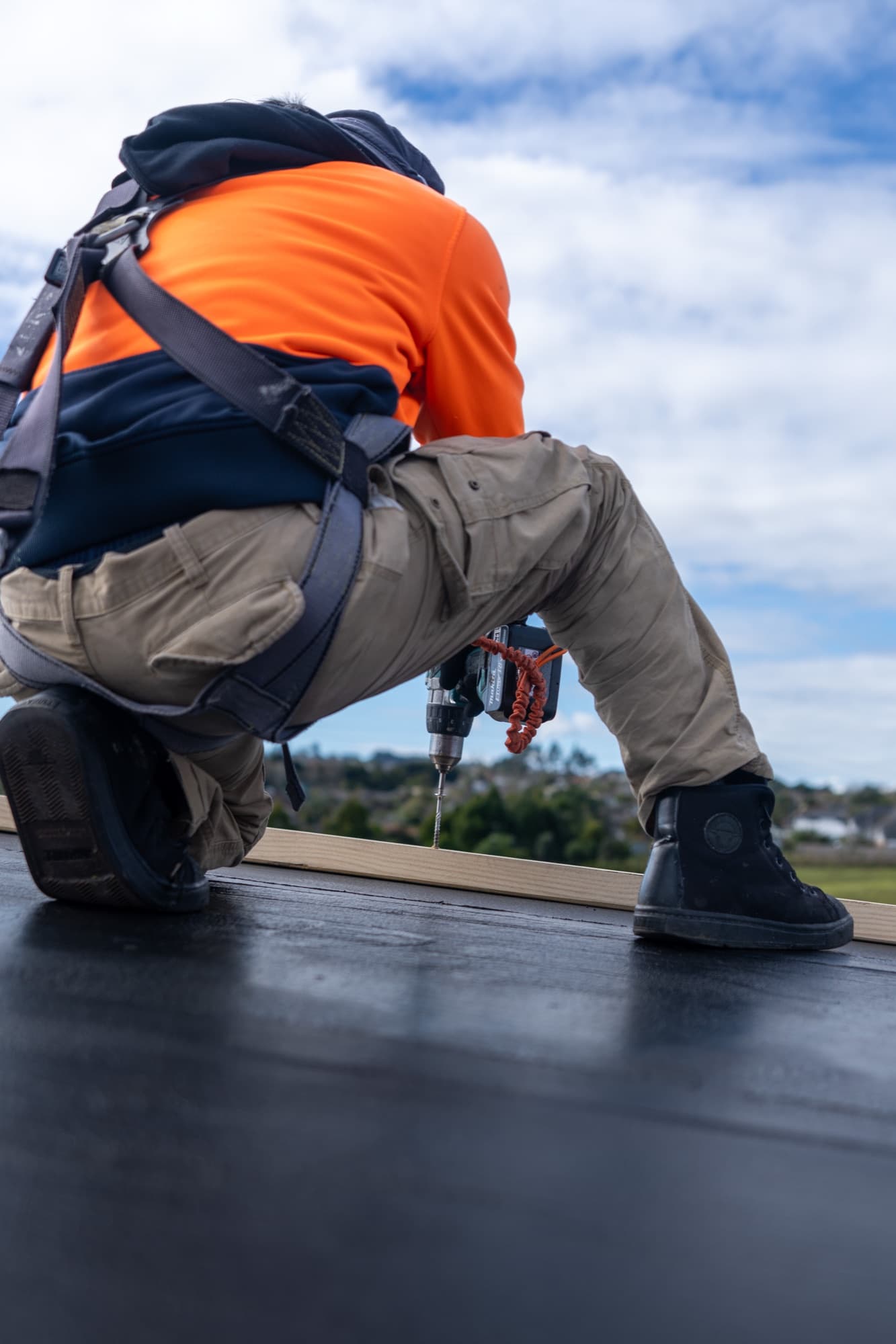 Roofer in a fall-arrest harness fixing a timber batten at the roof edge with a cordless drill