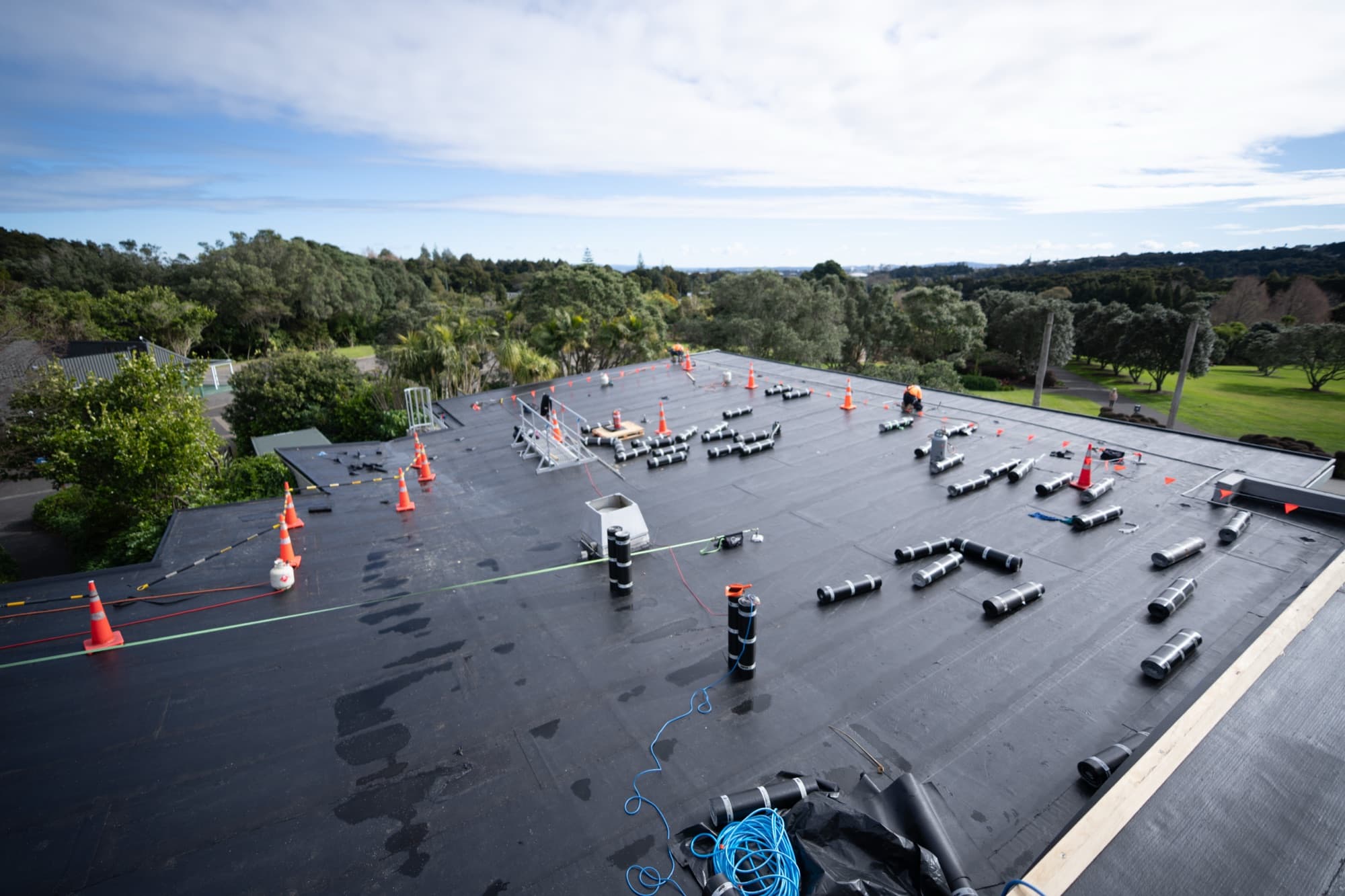 Wide view of the flat roof at Auckland Botanic Gardens Visitors Centre during remediation, with safety cones, edge delineation and membrane rolls staged across the deck