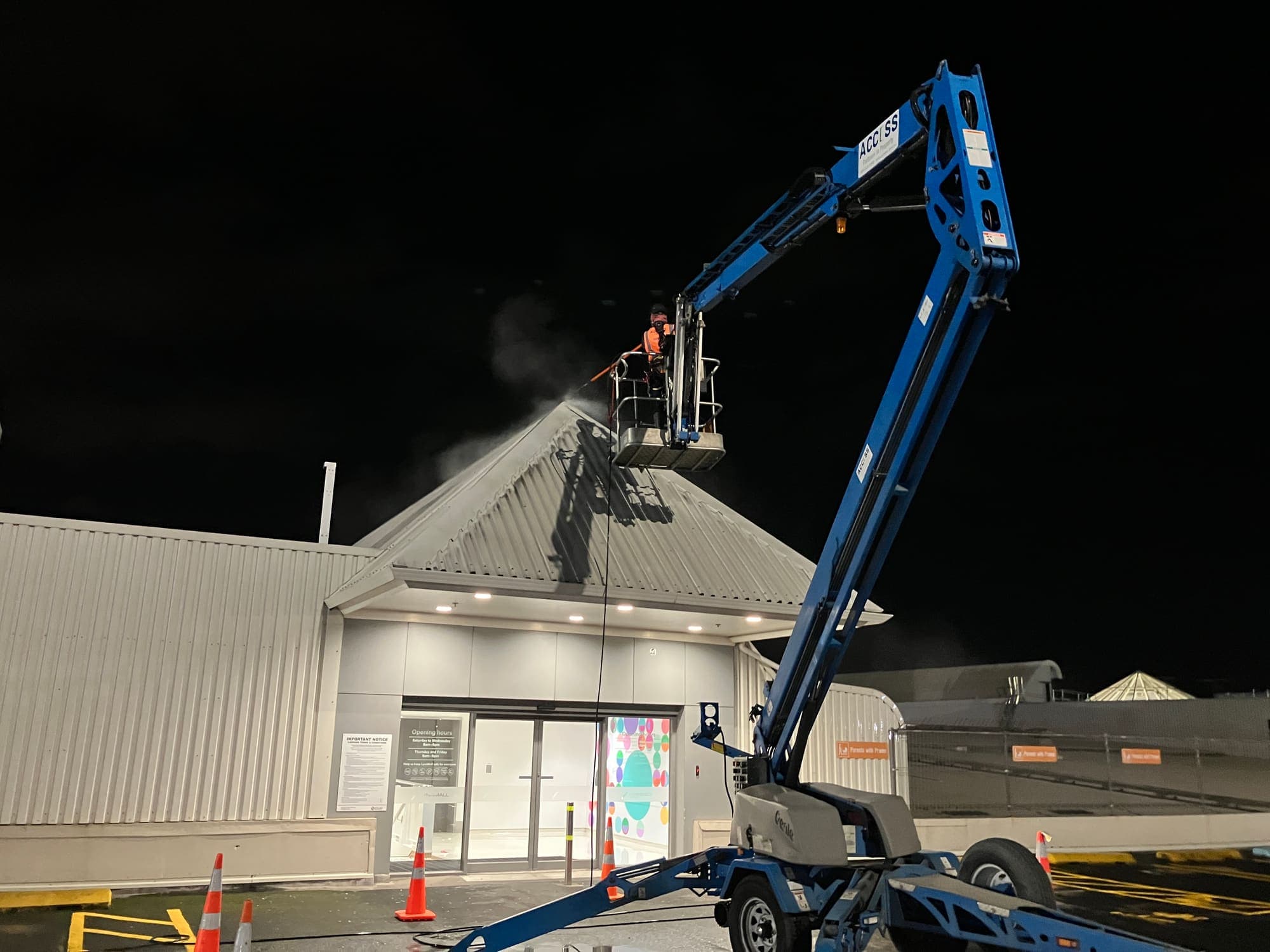 Technician spraying the LynnMall entrance canopy roof from an elevated work platform, wash mist visible against the night sky